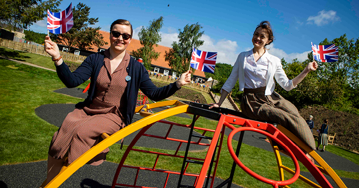 two women holding union jack flags smile at camera whilst sat on new playground at Beamish Museum